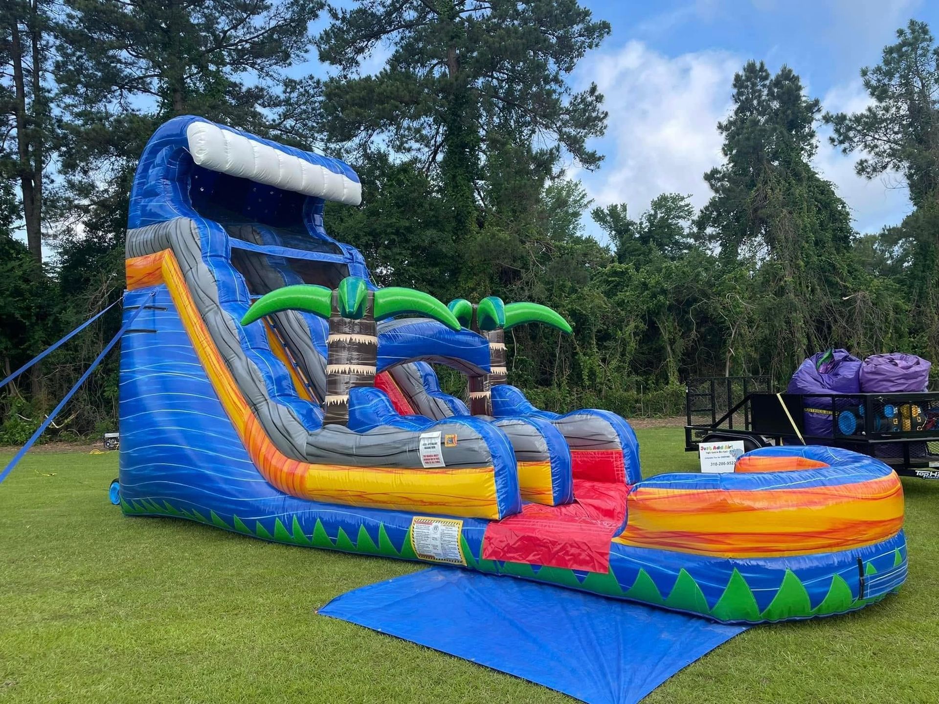 Colorful inflatable water slide and bounce house on grass at an outdoor park, with palm-tree decorations.