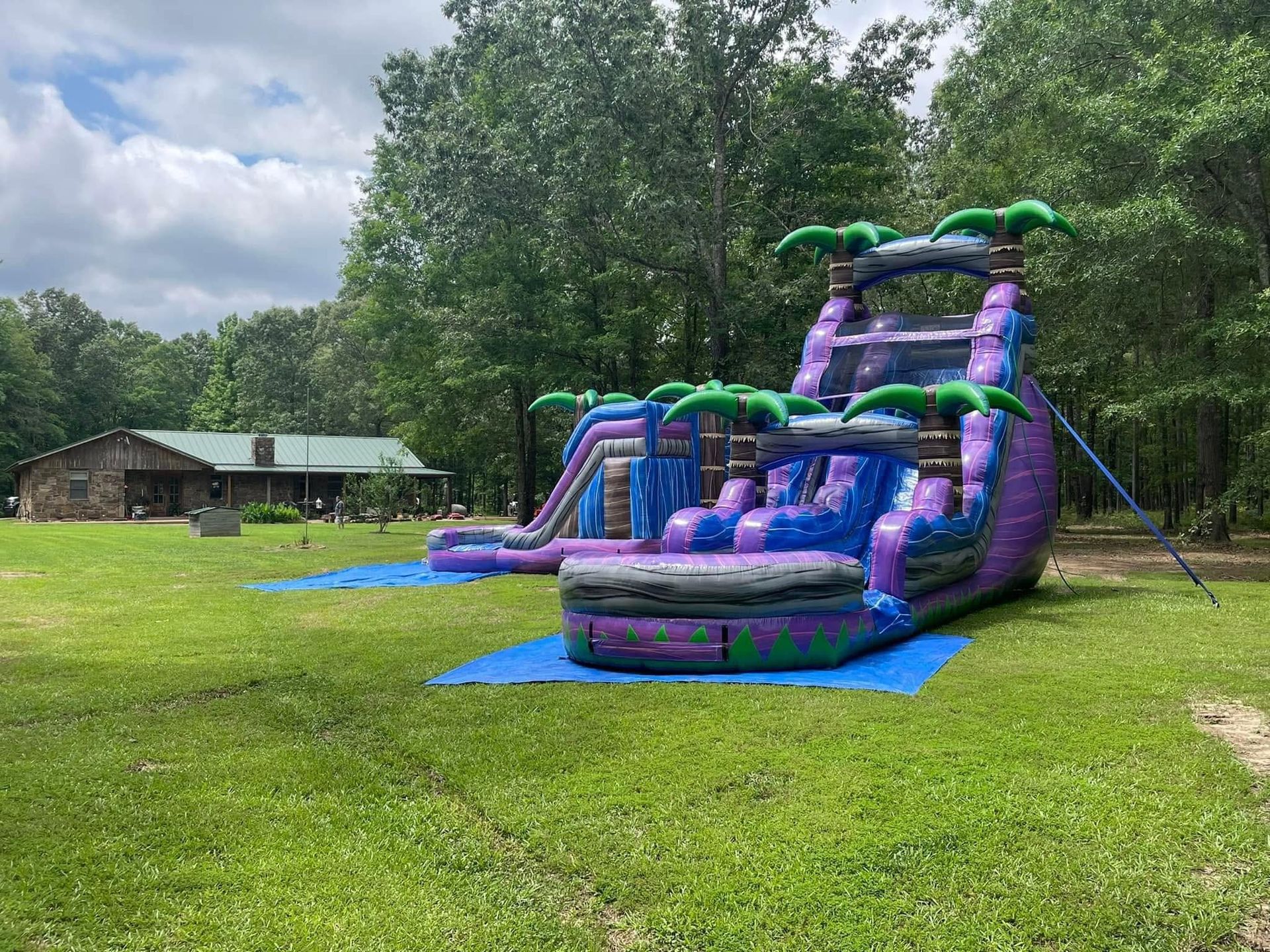 Purple-and-green inflatable water slide on a grassy lawn beside a house, with trees in the background