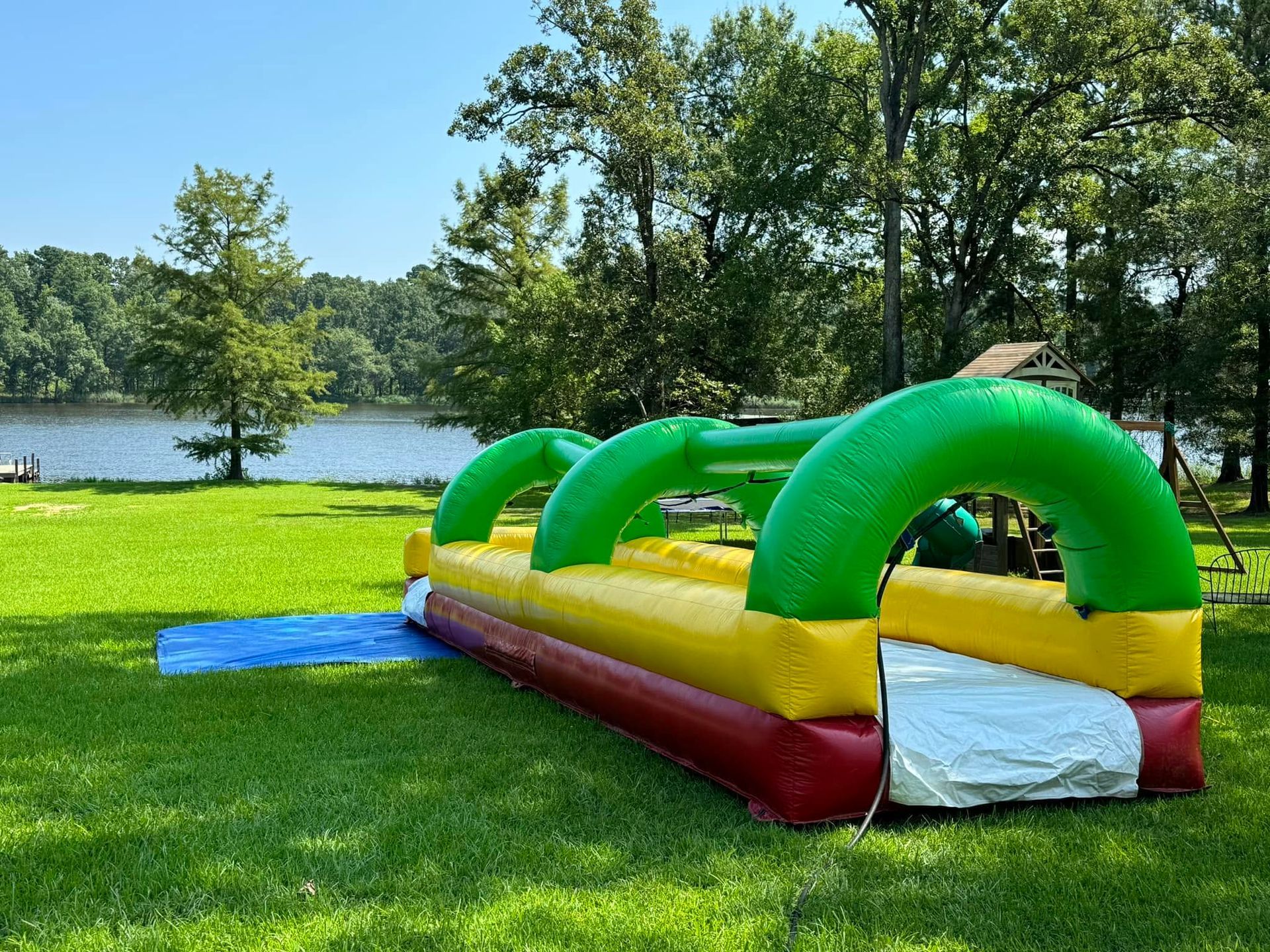 Colorful inflatable obstacle course on grass beside a lake, with trees and a bright blue sky