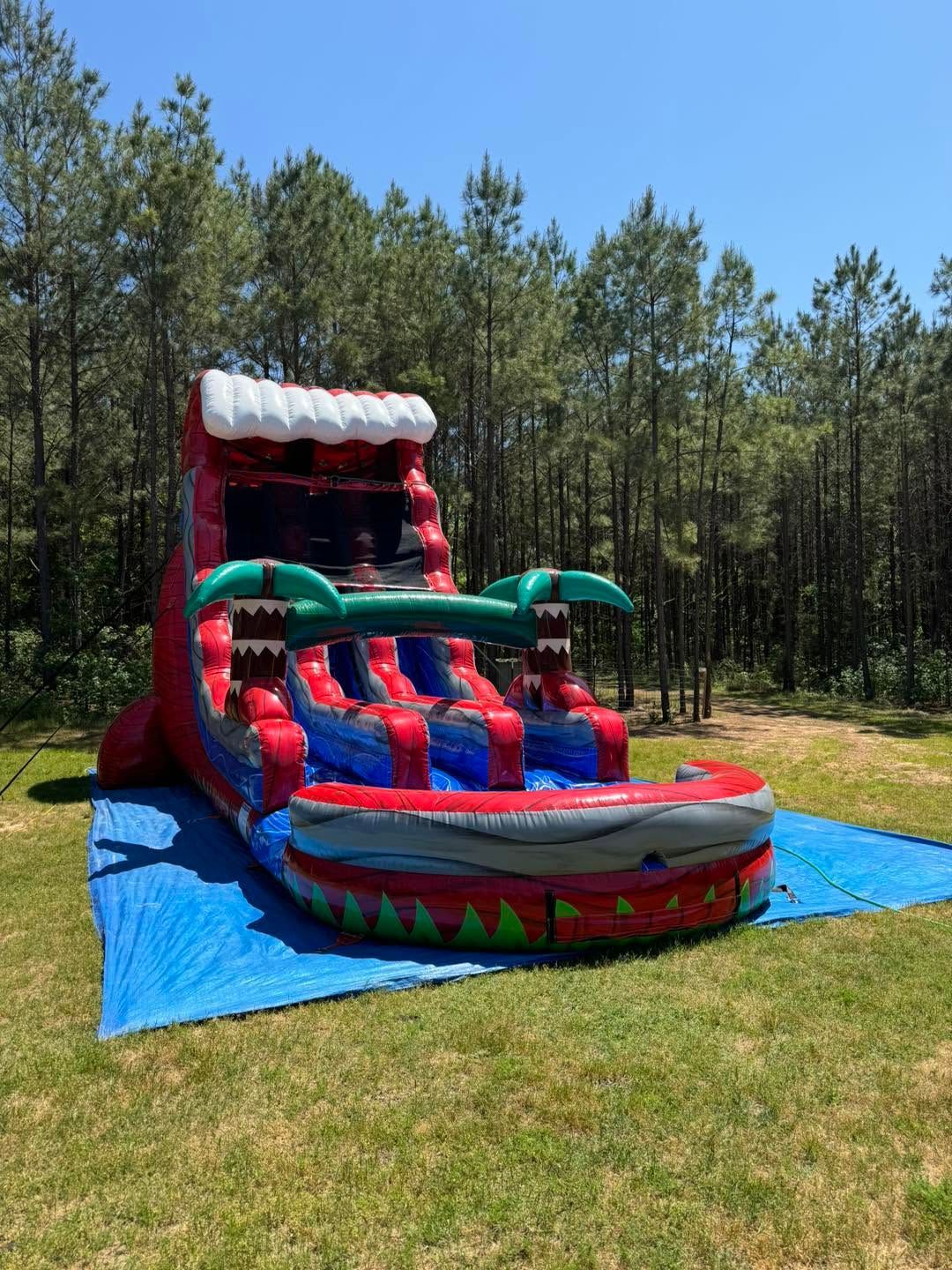 Inflatable red and teal bounce house slide on blue tarp in a grassy yard with pine trees.