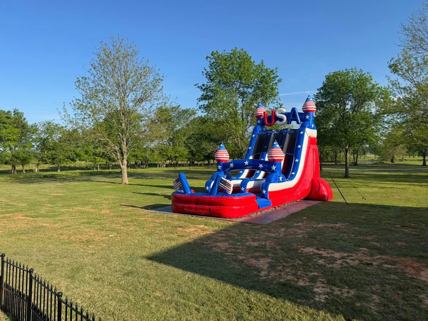 Red, white, and blue inflatable slide set up on a grassy field with trees and blue sky.