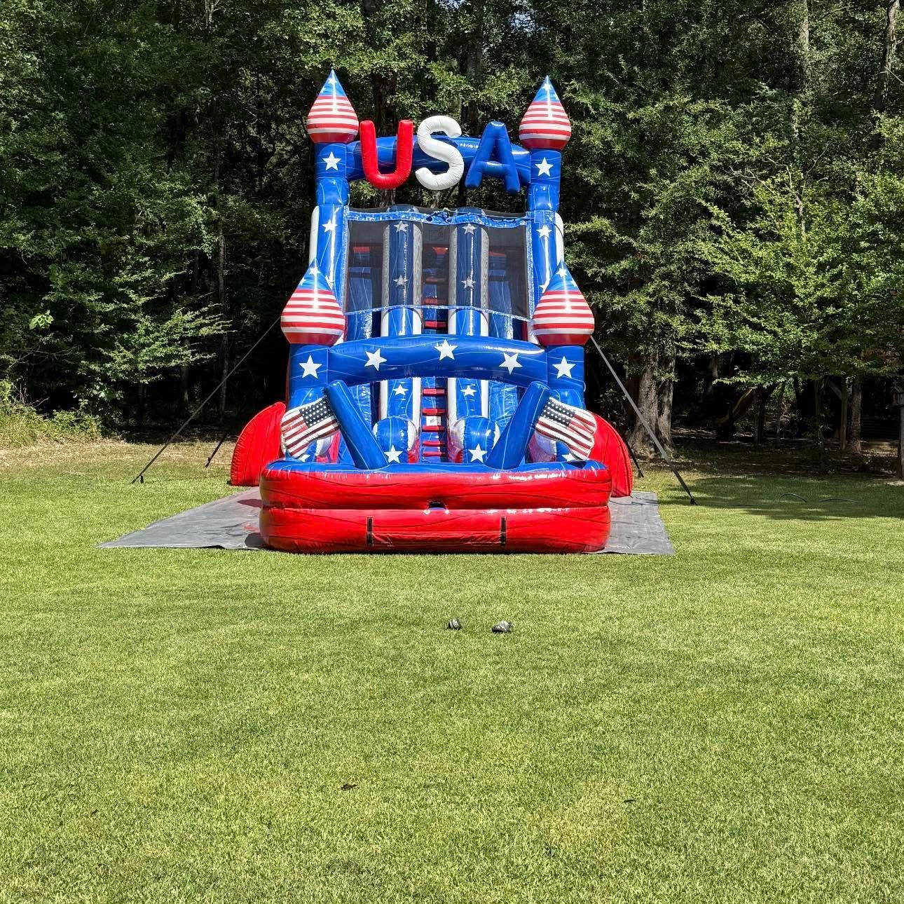 Red, white, and blue USA bounce house on grass in a wooded park