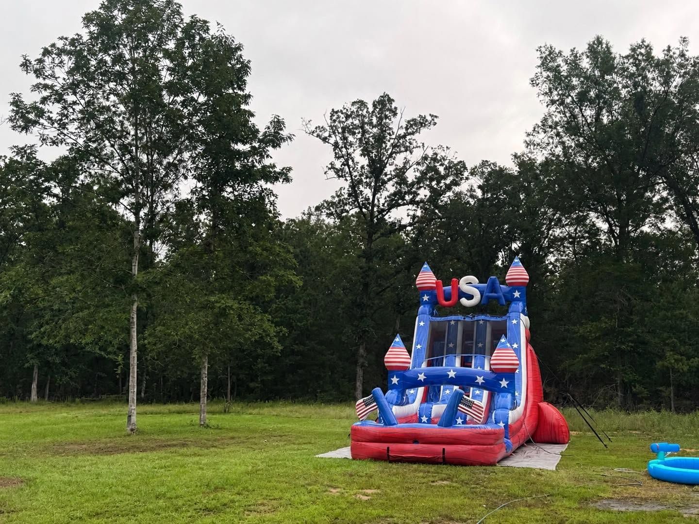 Red, white, and blue inflatable slide on a grassy field beside trees and an open lawn.