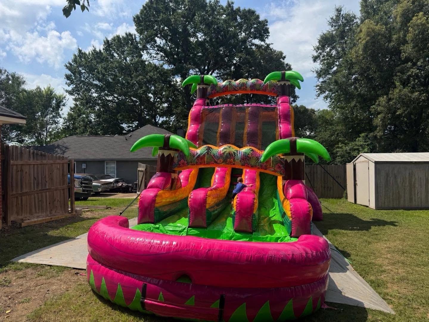 Bright pink and green inflatable water slide in a backyard with a house and trees under a blue sky