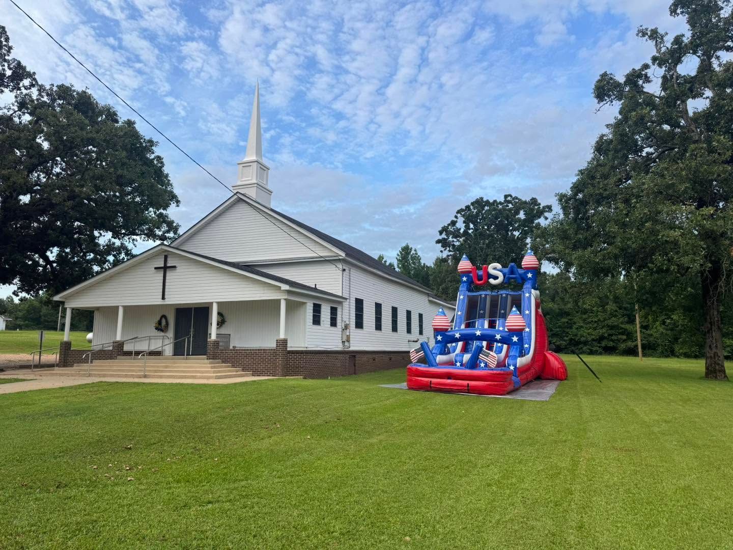 White church with steeple beside a red-and-blue inflatable bounce house on a grassy lawn