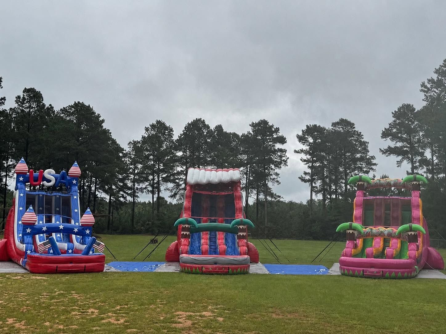 Three colorful inflatable bounce houses on a blue tarp in a grassy field under cloudy skies.