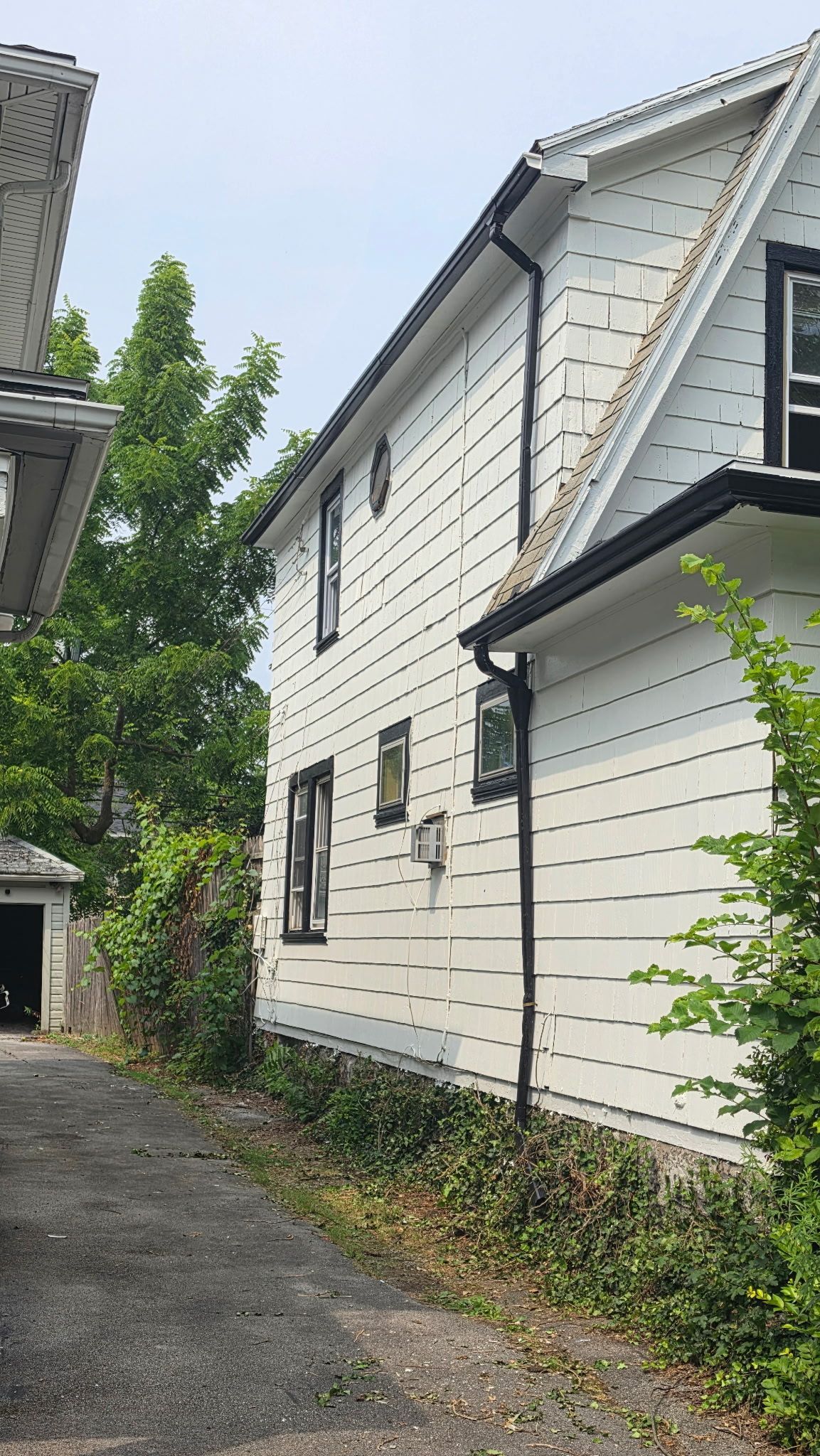White two-story house with black trim along a narrow driveway, viewed from the side.