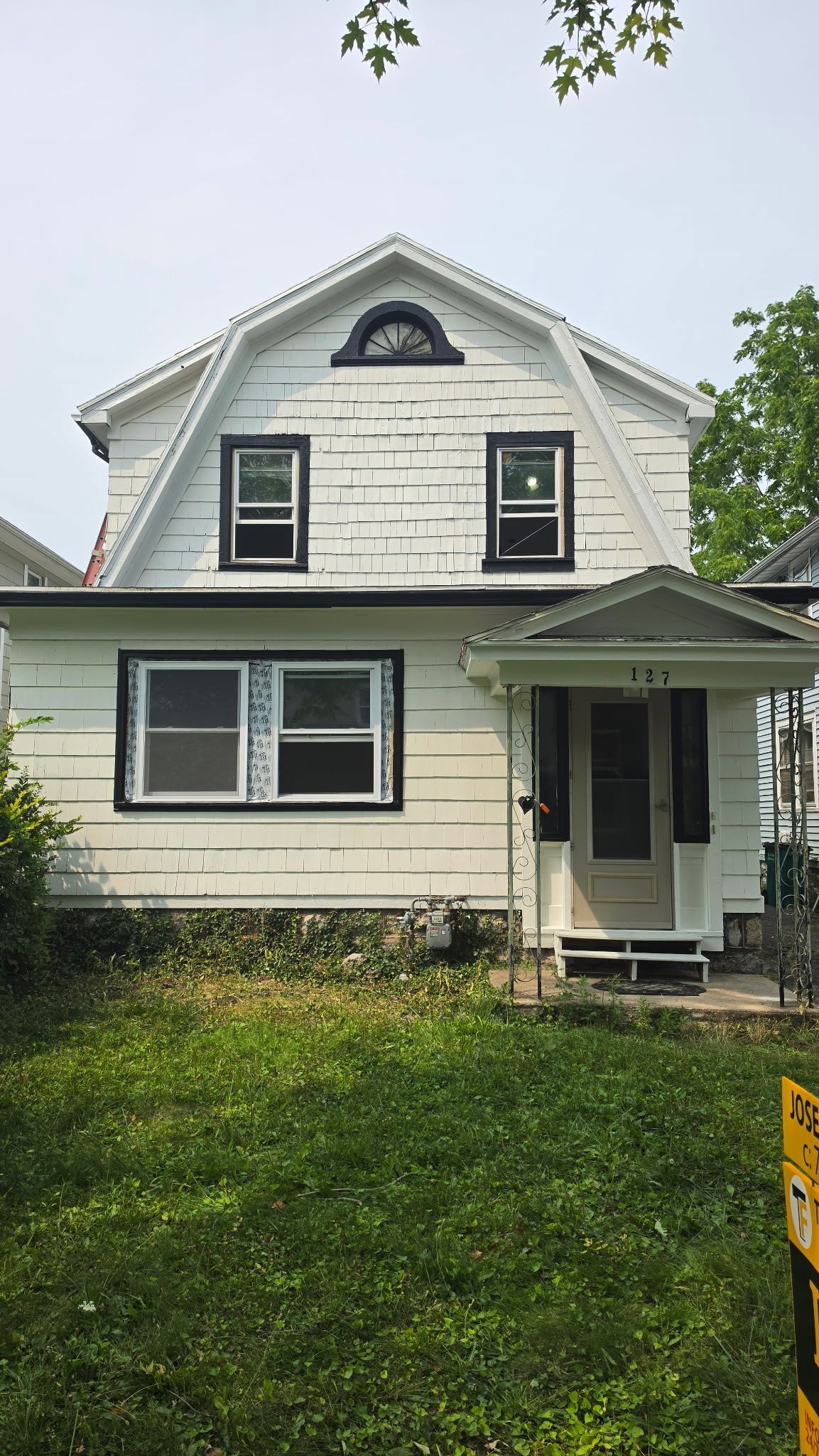 White two-story house with green trim, front porch, and overgrown lawn under a cloudy sky
