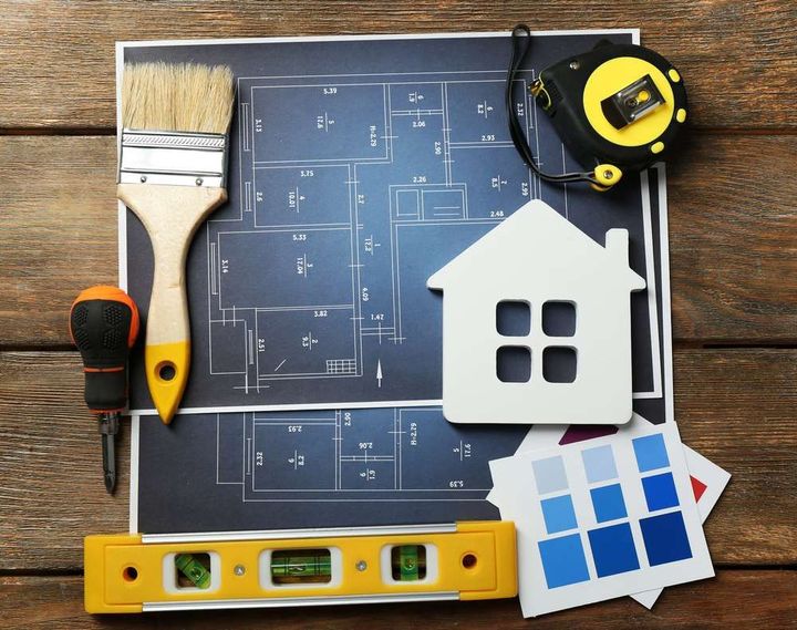 A wooden table topped with a blueprint , a brush , a tape measure , and a model house.