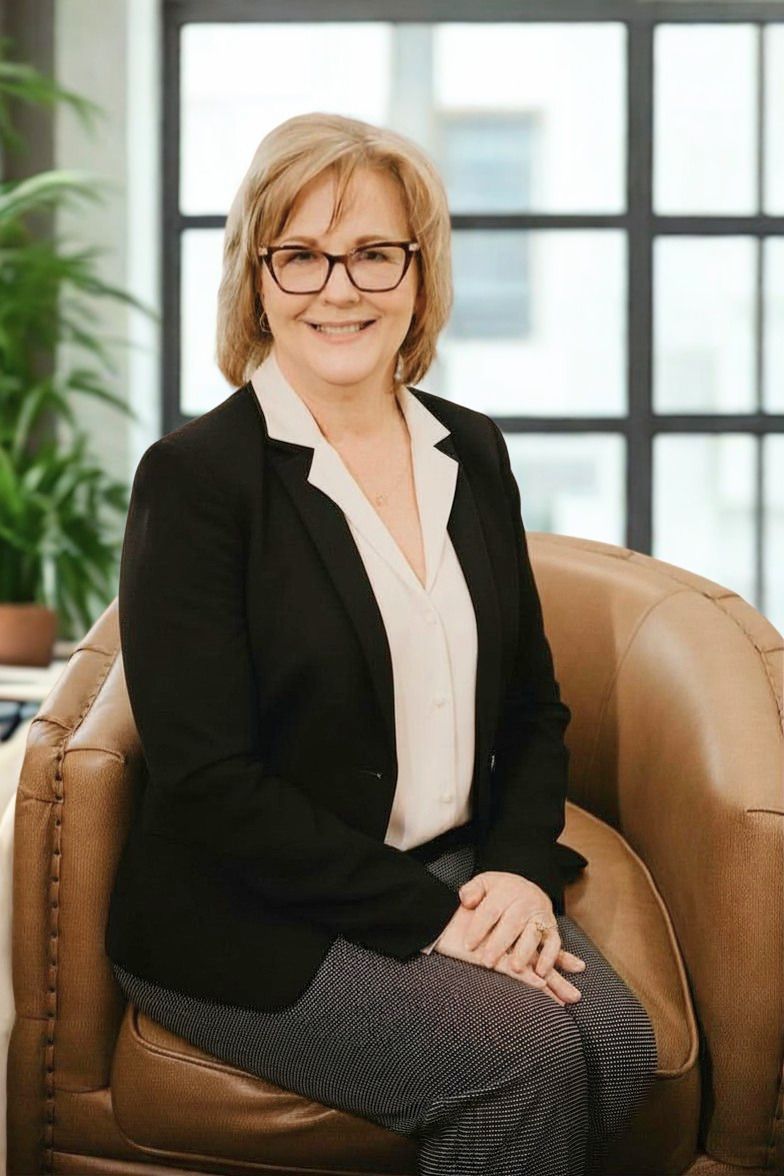 A woman wearing glasses and a grey jacket is smiling with her arms crossed