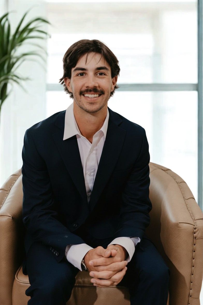 A smiling person with dark hair and a mustache wearing a dark suit and white collared shirt, seated in a beige chair.