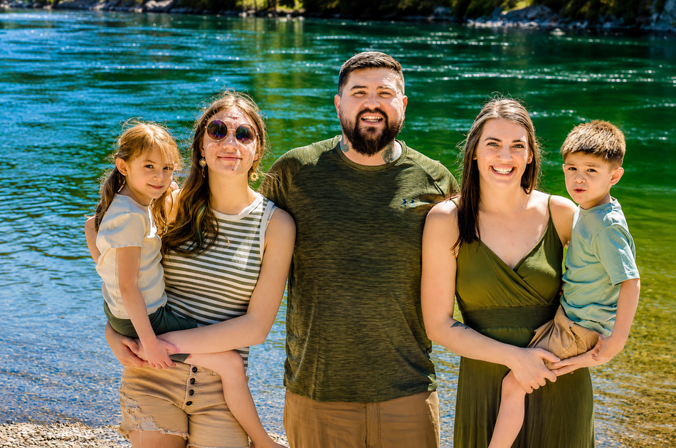 Family of four standing together outdoors near water, smiling at the camera.