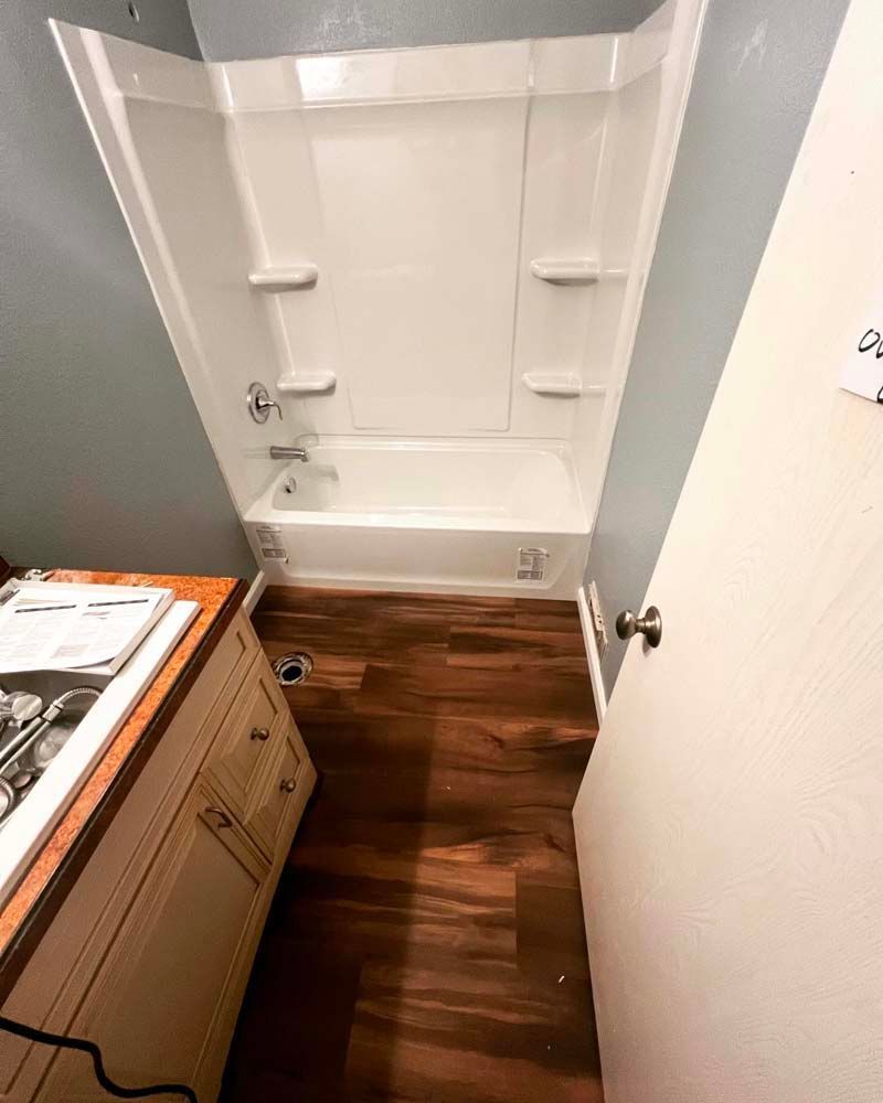 Small bathroom with a white bathtub-shower, wood-look floor, sink cabinet, and open door.