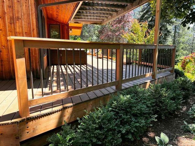 Wooden deck with railing beside a house, overlooking a garden with shrubs and trees.