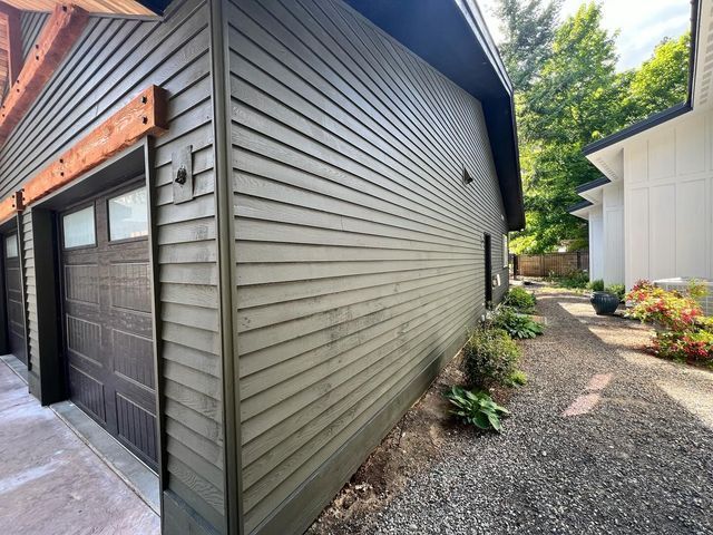 Side of a modern house with gray siding, garage doors, and a gravel walkway beside a white outbuilding.