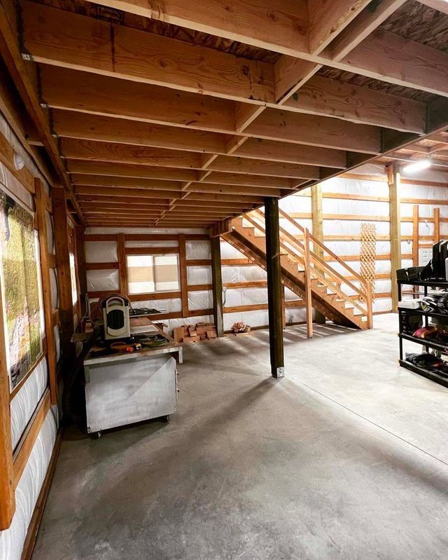 Unfinished basement with exposed wooden beams, concrete floor, staircase, and storage shelves.