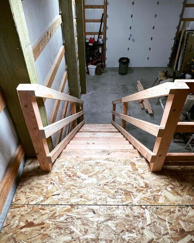 Wooden staircase under construction with railings, leading down to a basement floor with tools and a bucket.