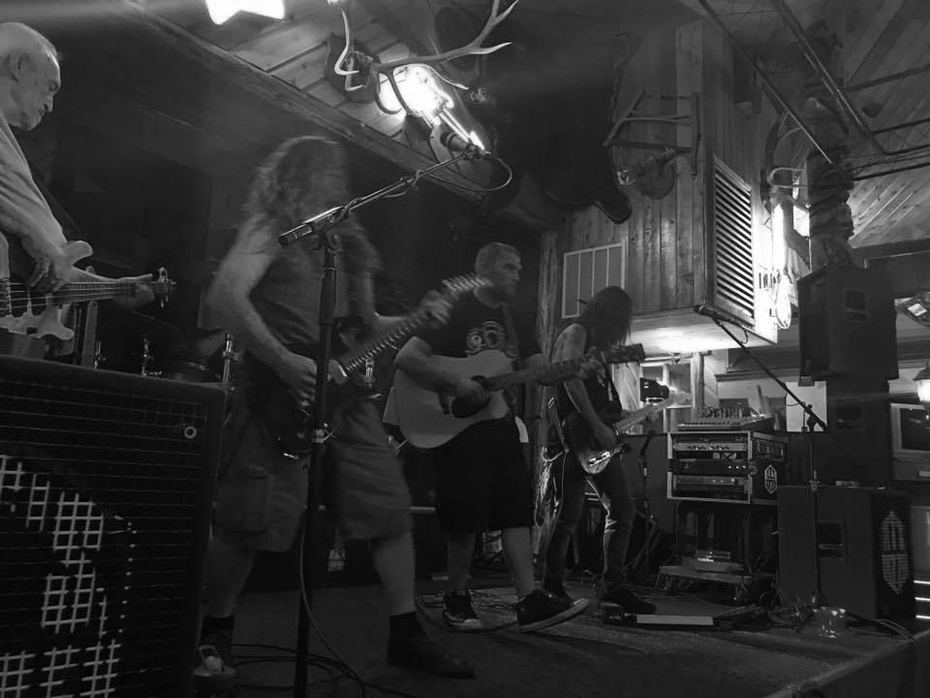 Band performing on stage in a rustic venue, playing guitars, and bass. Black and white.