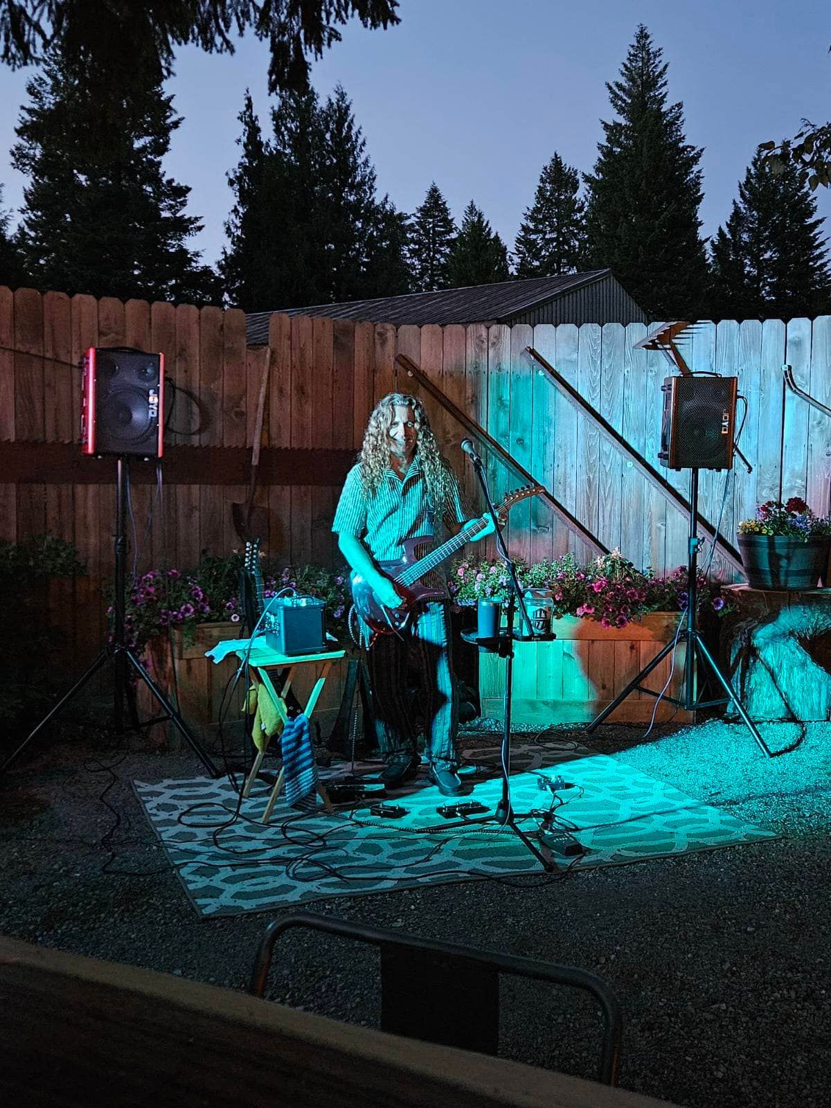 Musician playing guitar on an outdoor stage lit blue, with drums and speakers at dusk
