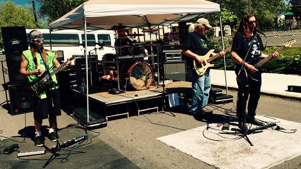 Four musicians play guitars and drums outdoors under a canopy at a small stage setup.