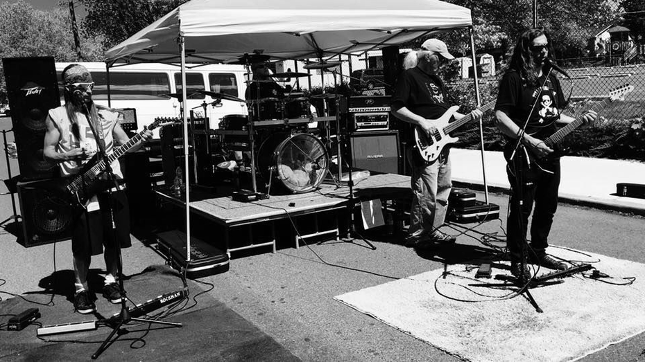 Band playing music outdoors under a canopy. One plays drums, others guitar and bass. Black and white photo.