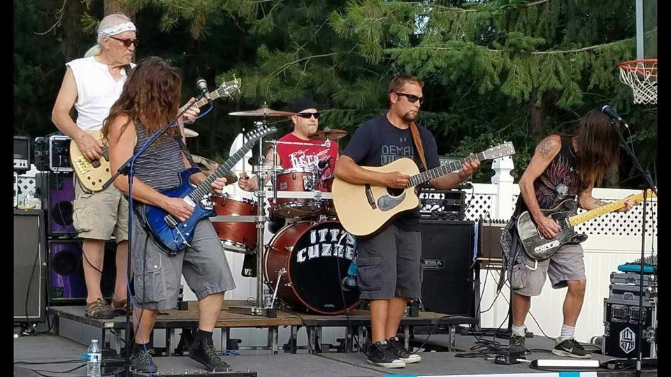 A band performs on an outdoor stage, featuring a drummer and musicians playing electric and acoustic guitars.