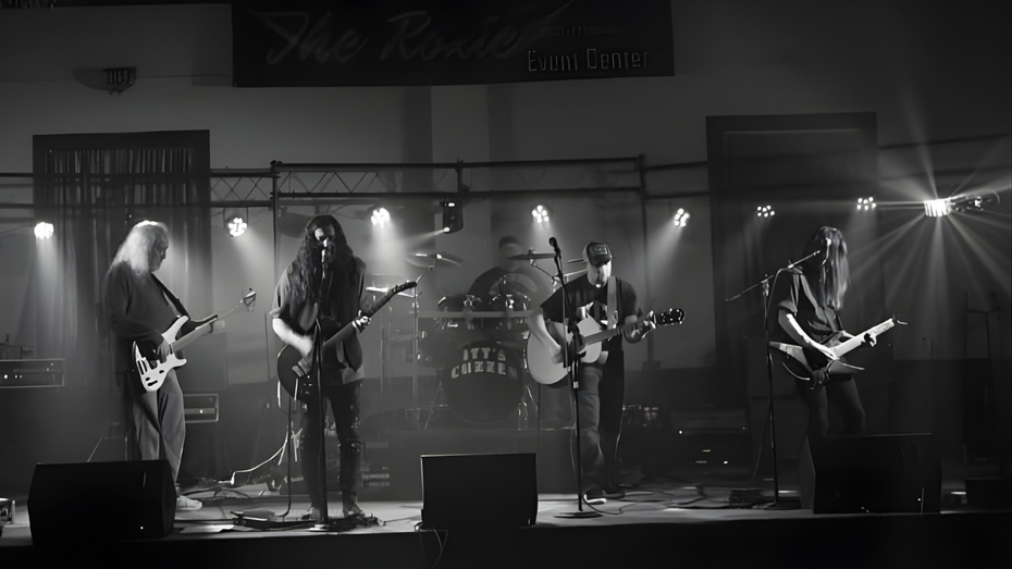 Band performing live on stage under spotlights. Musicians with guitars, drums visible. Black and white.