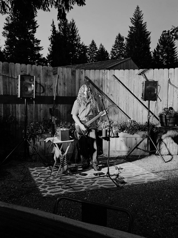 Musician playing electric guitar in a backyard, with speakers and microphone set up near a fence.