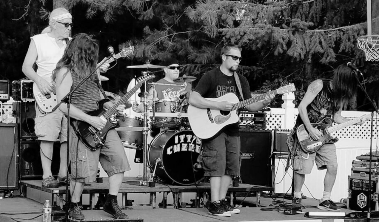 Band performing outdoors, on stage, in black and white. Musicians playing guitar, bass, and drums.