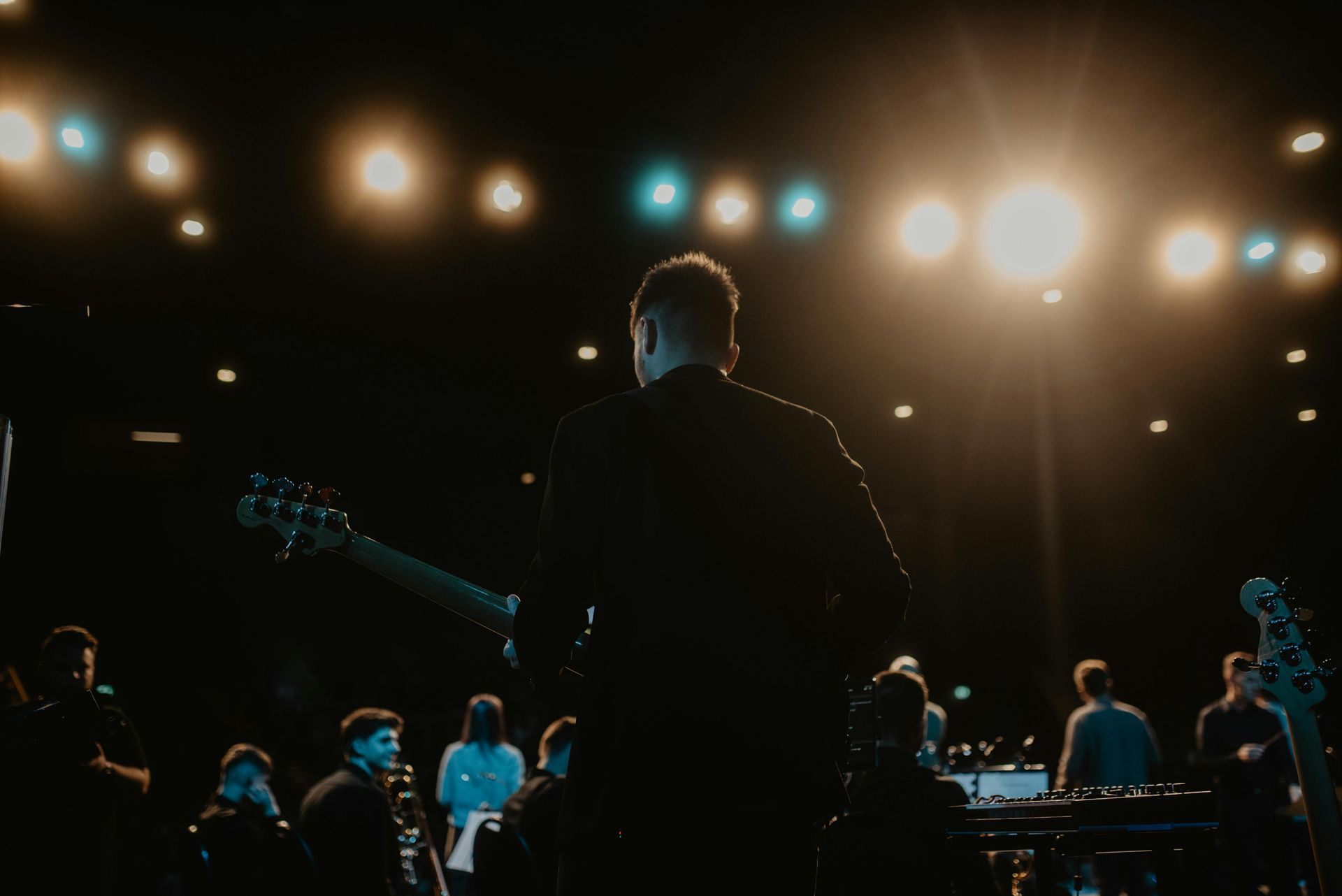Musician on stage seen from behind, facing band and bright concert lights in a dark venue