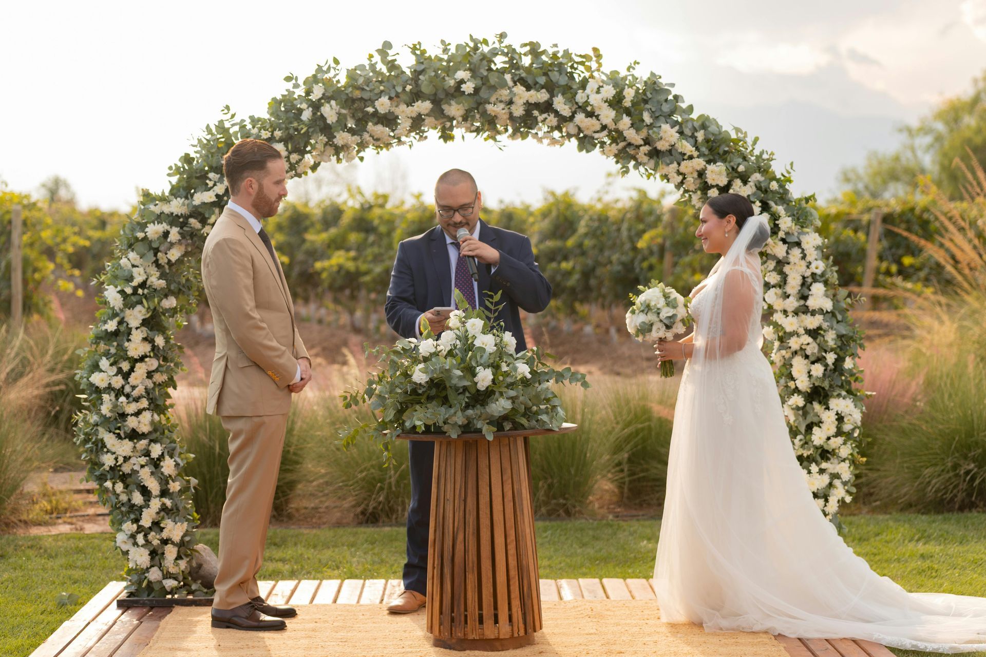 A couple stands under a circular floral arch in a vineyard during their wedding ceremony as an officiant reads aloud.