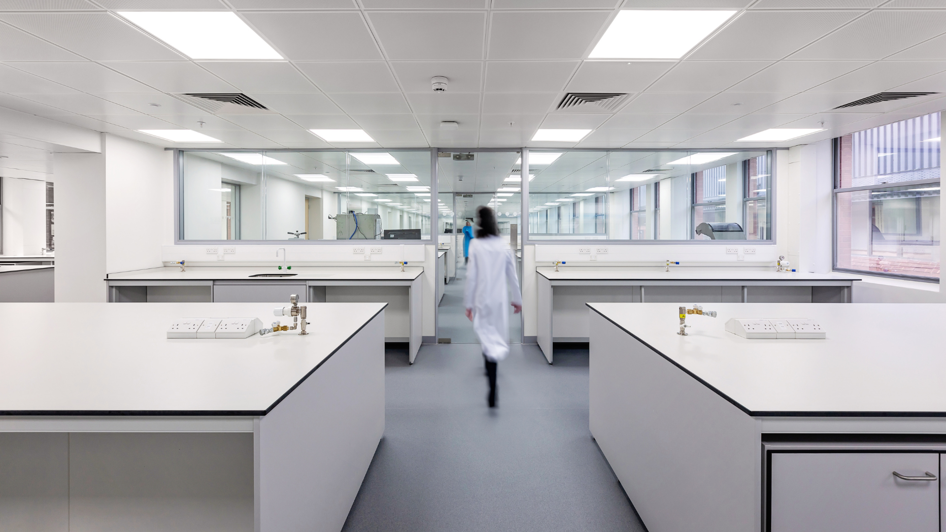 Bright laboratory with white benches and a person in a lab coat walking away down the aisle