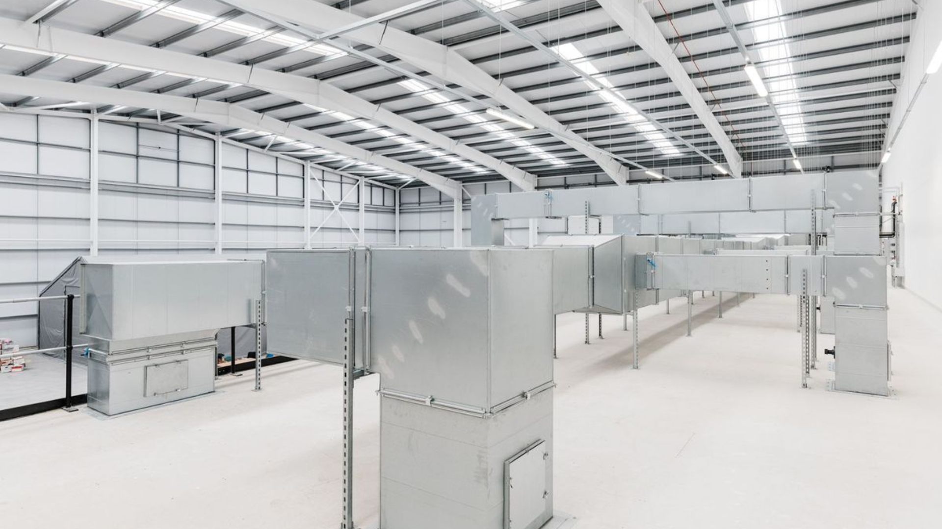 Bright empty warehouse interior with white floors, gray equipment, and high metal ceiling beams