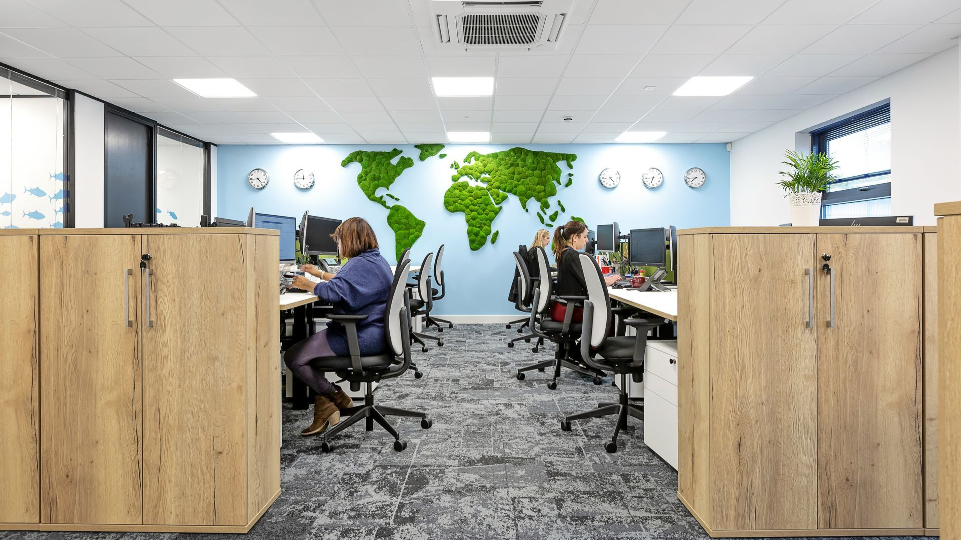 Modern open office with workers at desks, wood dividers, and a green world map mural on the wall