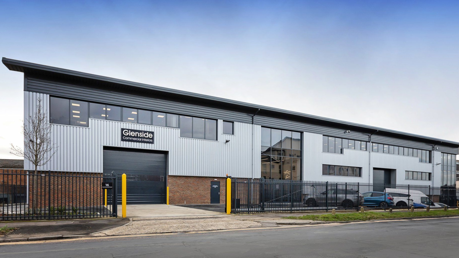 Modern industrial building with black fence, yellow bollards, and cloudy sky