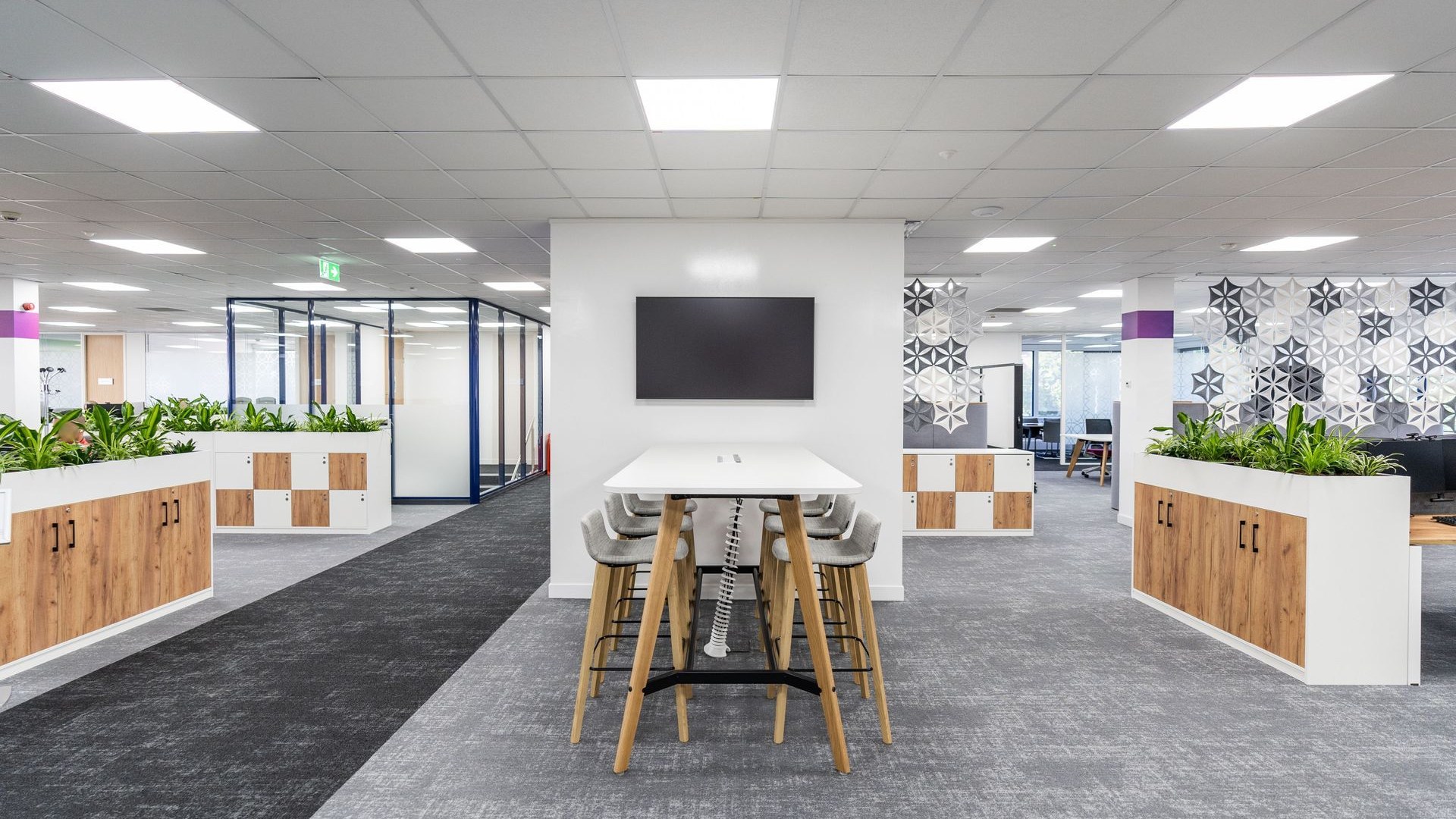 Modern office interior with desks, chairs, white walls, and a wall-mounted TV in the center.