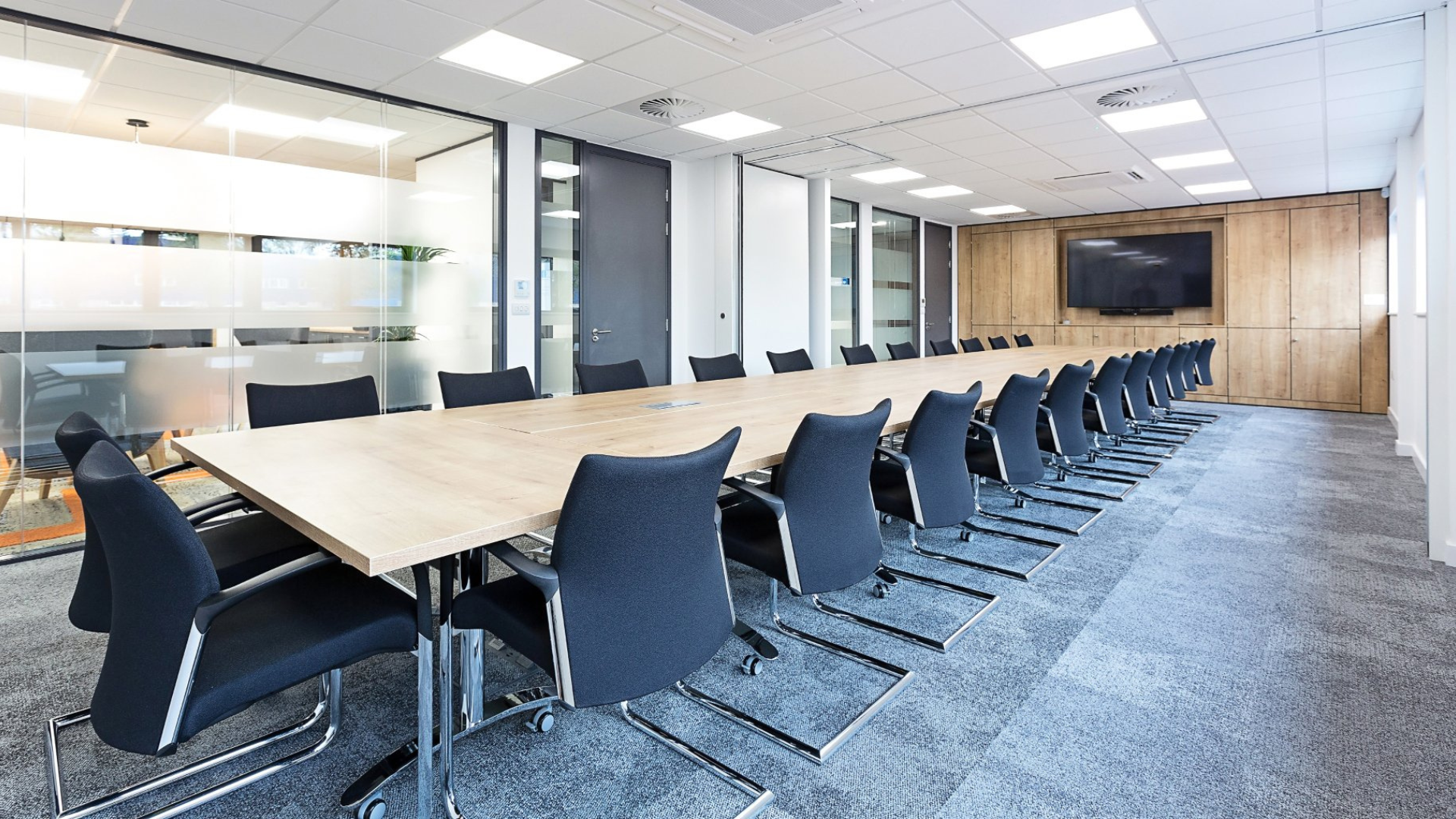 Modern conference room with a long wooden table, black chairs, glass walls, and carpeted floor