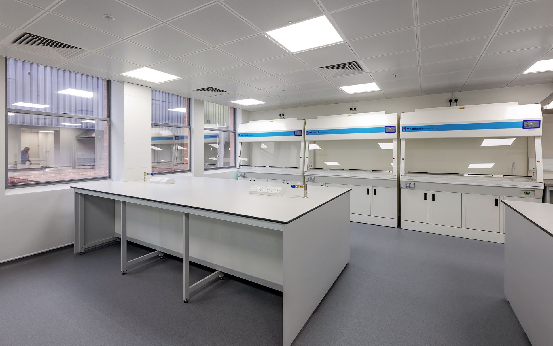 Bright laboratory with white workbenches, cabinets, and large windows under fluorescent ceiling lights.