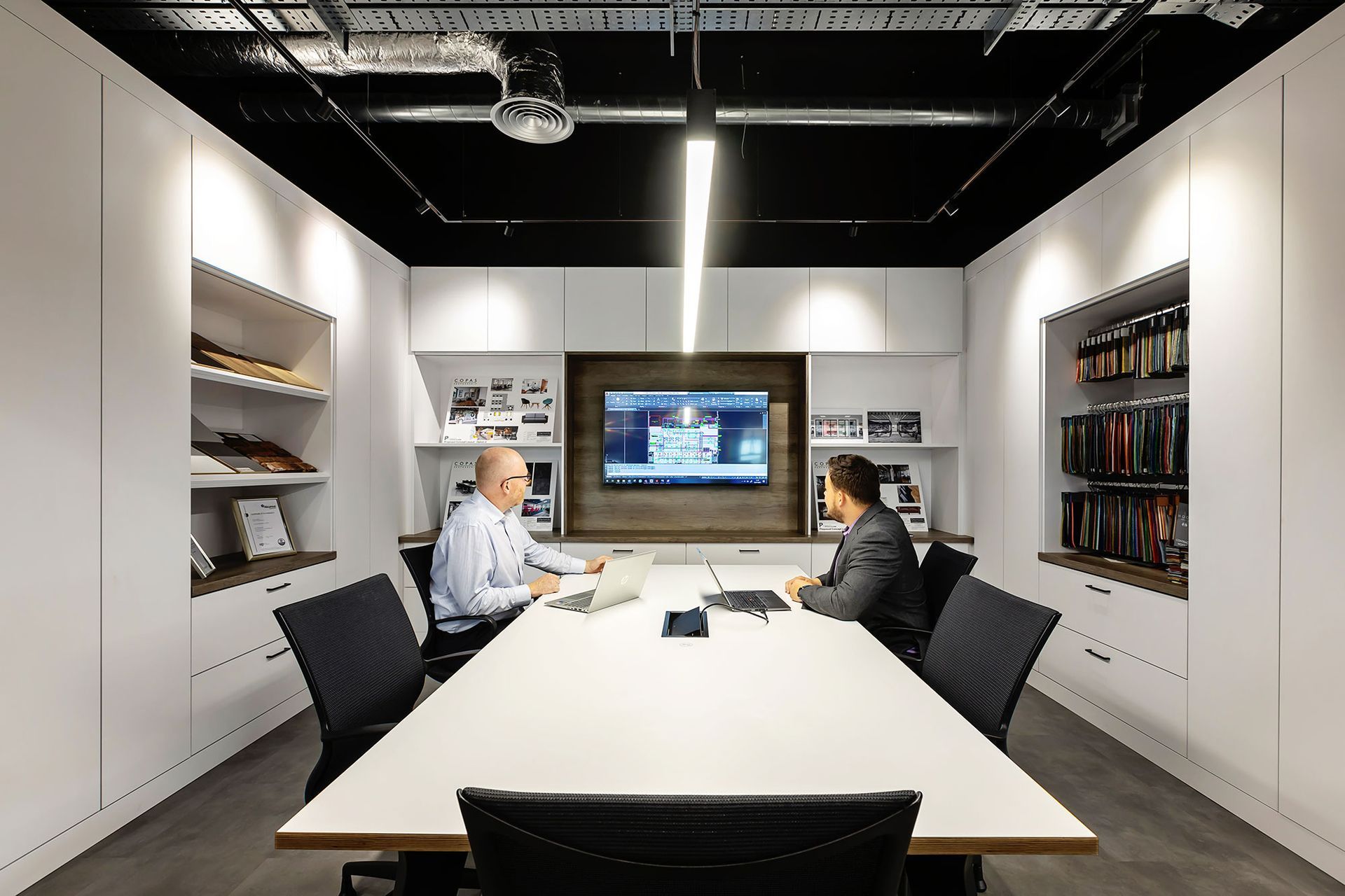 Modern conference room with two people seated at a long white table facing a wall-mounted screen.