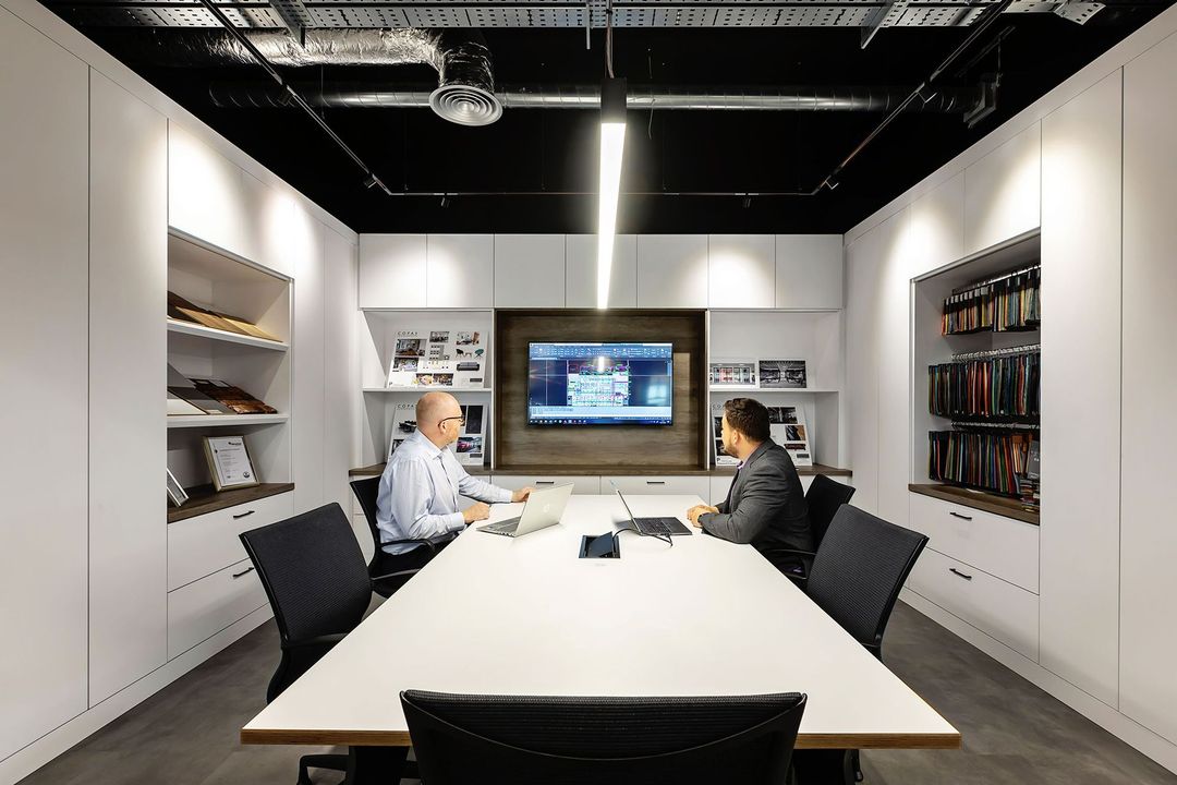 Modern conference room with two people seated at a long white table facing a wall-mounted screen.