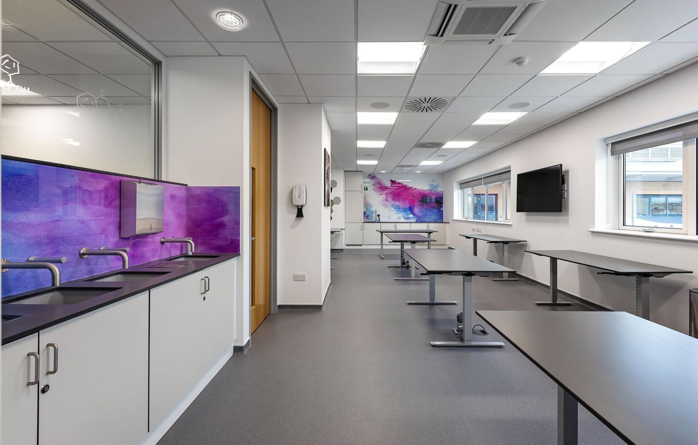Modern classroom or lab with white cabinets, purple backsplash, tables, wall-mounted screens, and bright windows