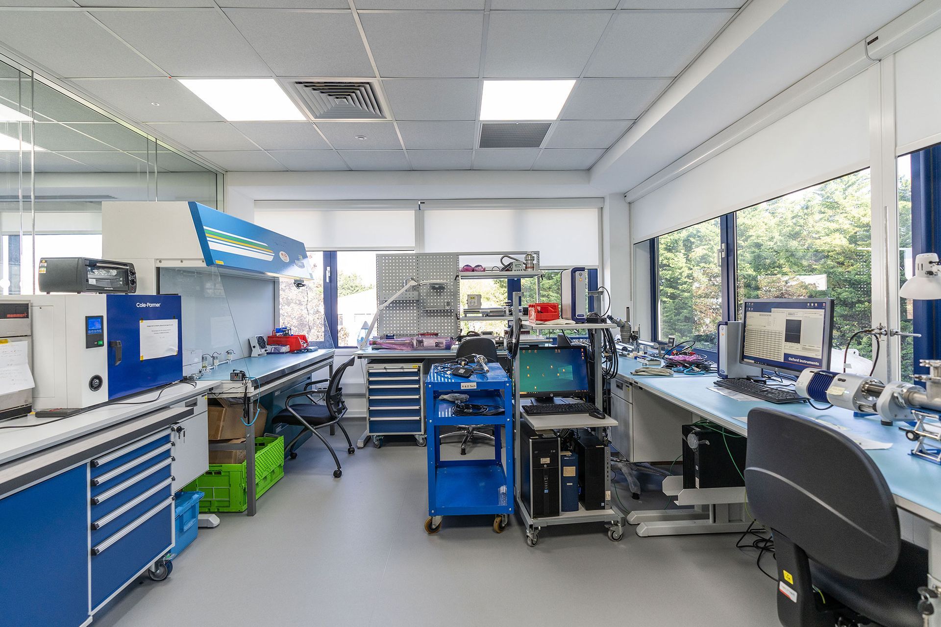 Bright electronics lab with blue workbenches, equipment, and a windowed office area