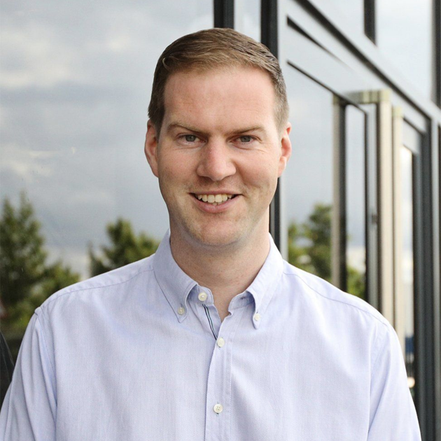 Smiling man in a light blue shirt standing outdoors beside a modern building