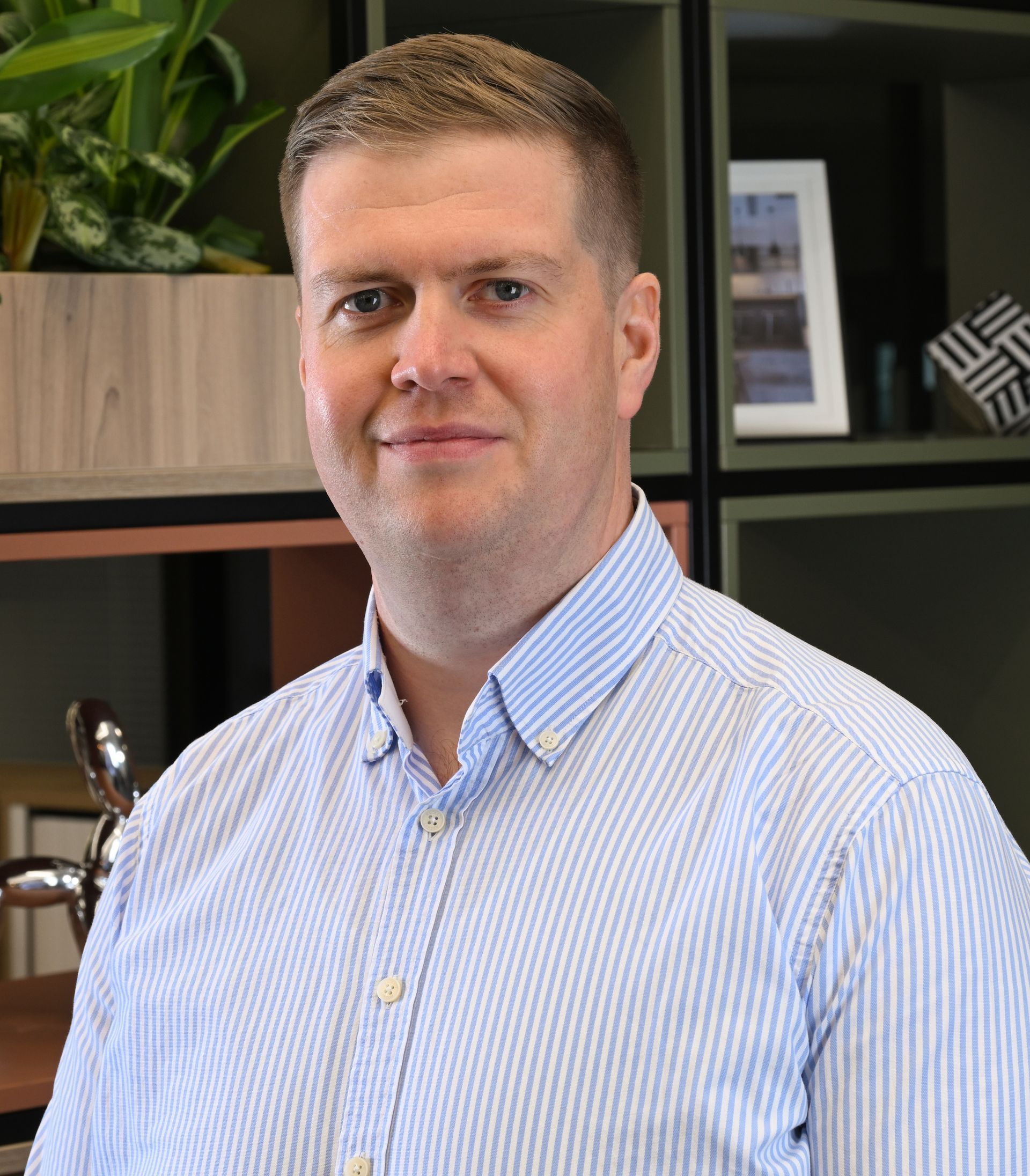 Portrait of a man in a light blue shirt standing indoors against a leafy wall and wood slats