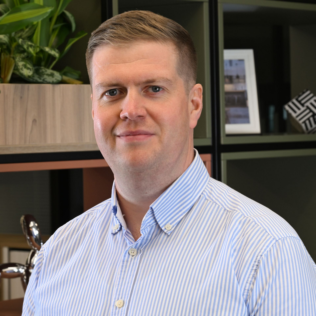 Portrait of a man in a light striped shirt standing indoors against a green plant and wood-paneled background