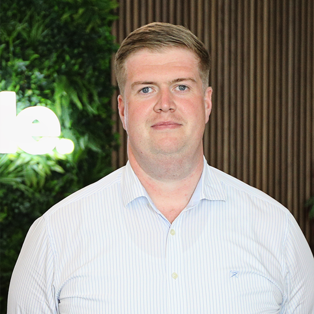 Portrait of a man in a light striped shirt standing indoors against a green plant and wood-paneled background