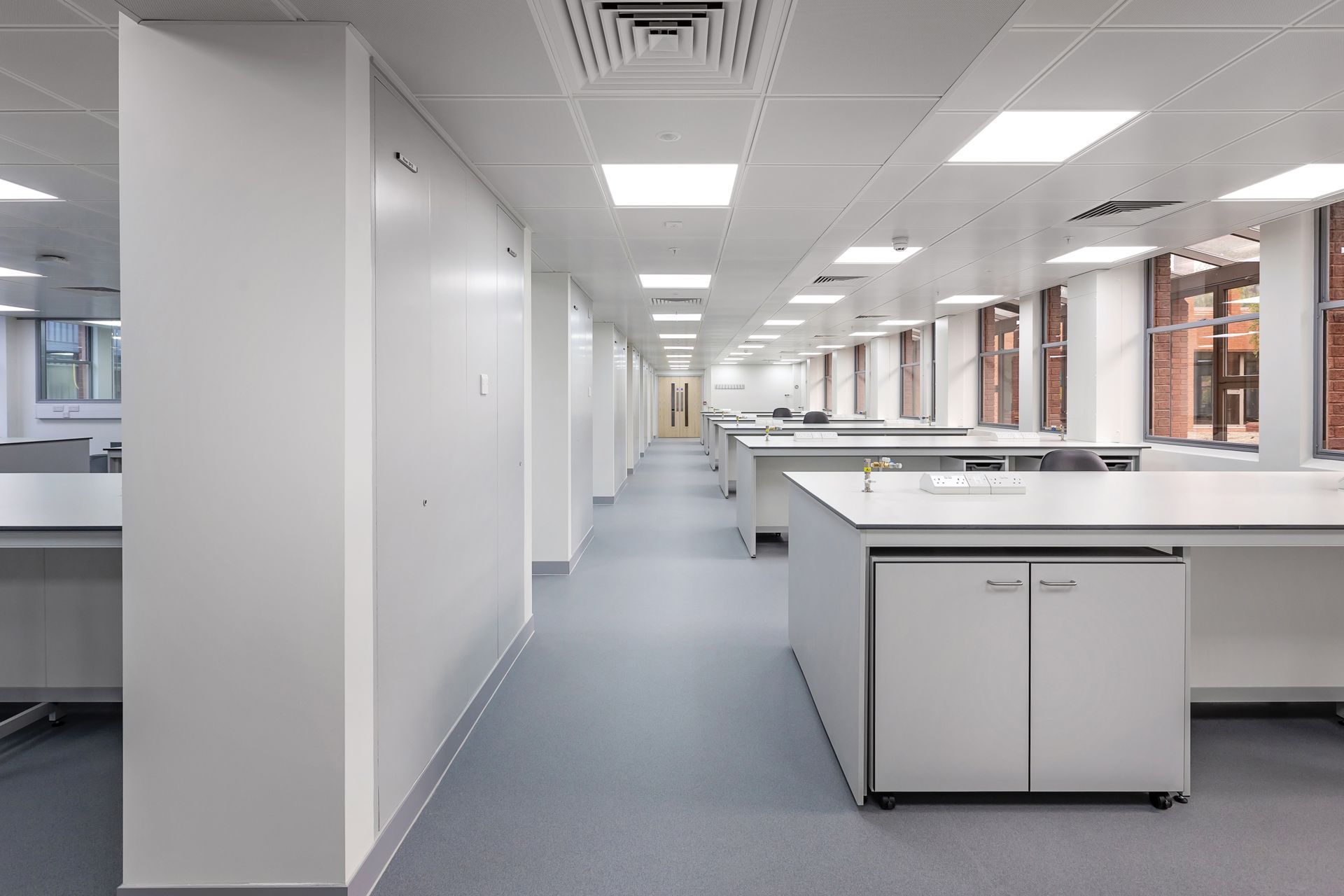 Empty modern office corridor with white walls, gray carpet, desks, and large windows.