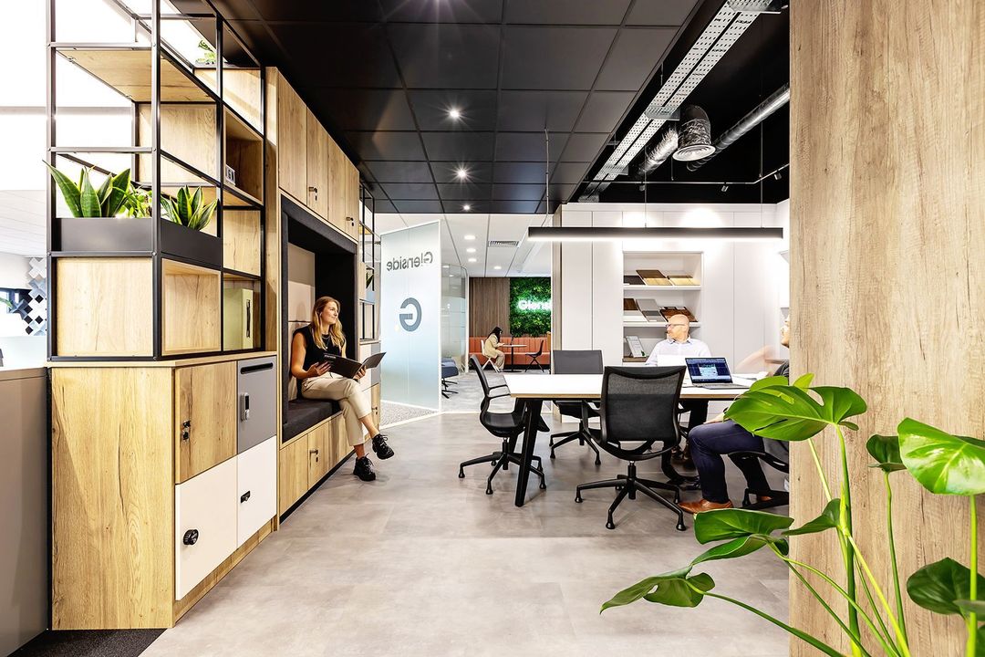 Modern office workspace with desks, shelves, plants, and two people meeting in a bright open area