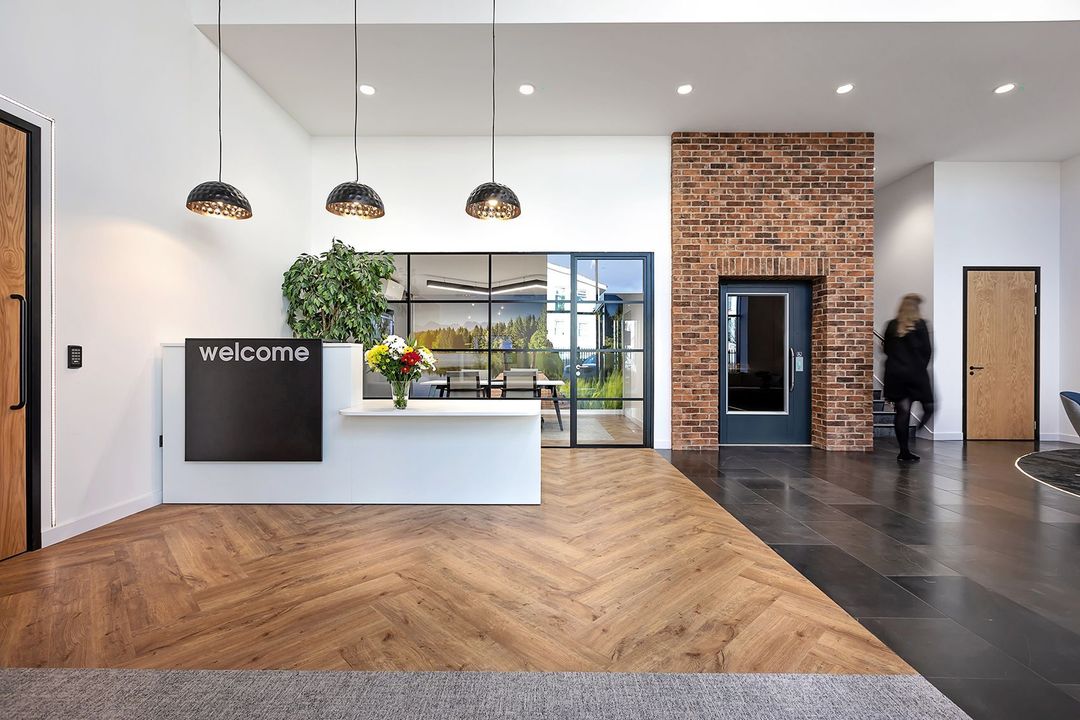 Modern lobby with reception desk, hanging lights, brick elevator wall, and wooden floor