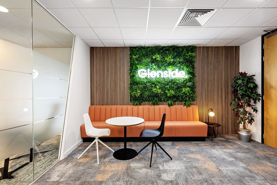 Modern office lounge with orange sofa, white round table, chairs, plants, and illuminated greenery wall.